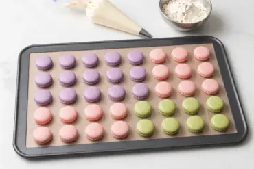 Silicone macaron baking mat with printed template circle guides and piped pastel macaron batter on a marble counter.